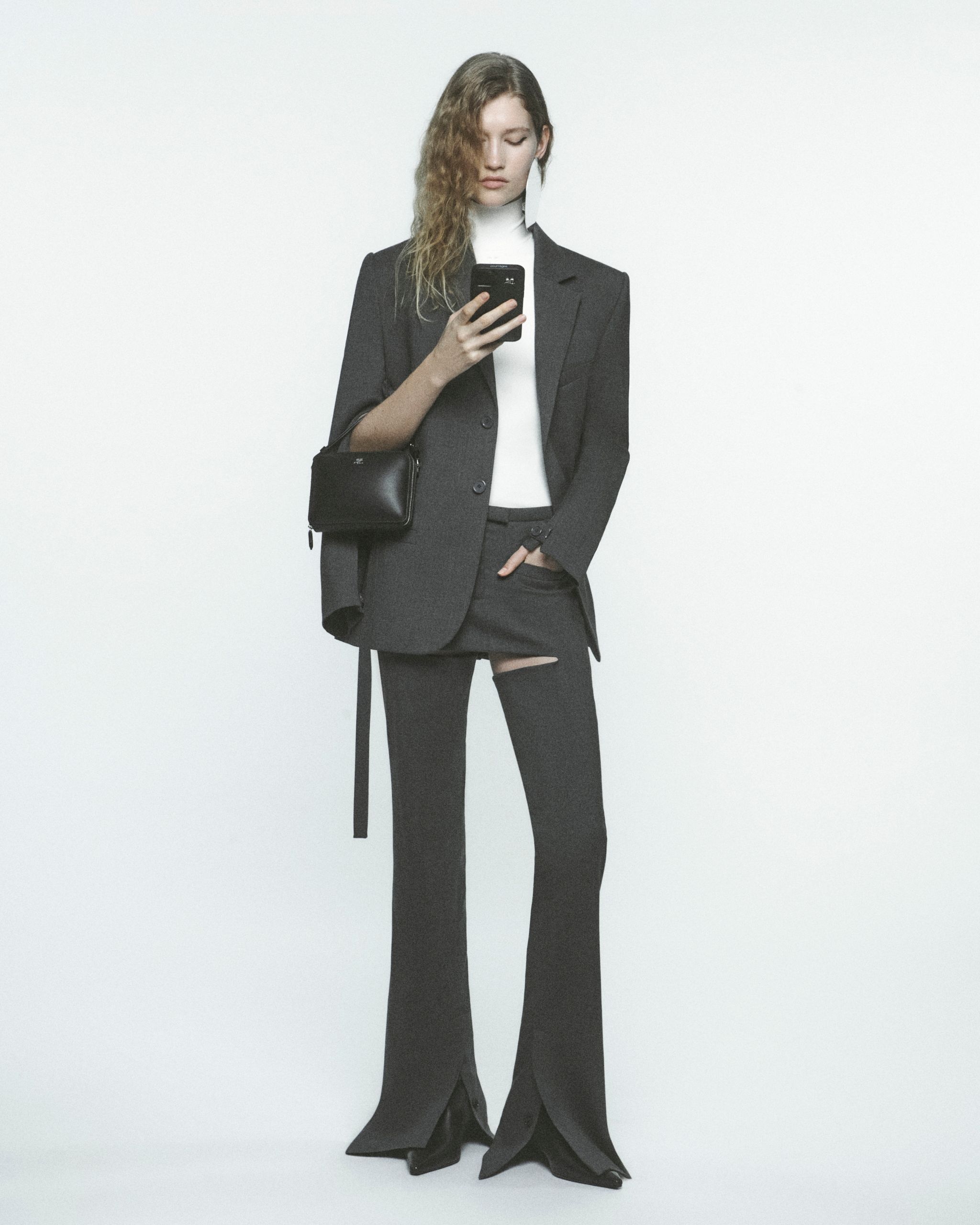 Femme élégante en costume noir et pantalon déchiré, tenant un smartphone, représentant Courrèges.