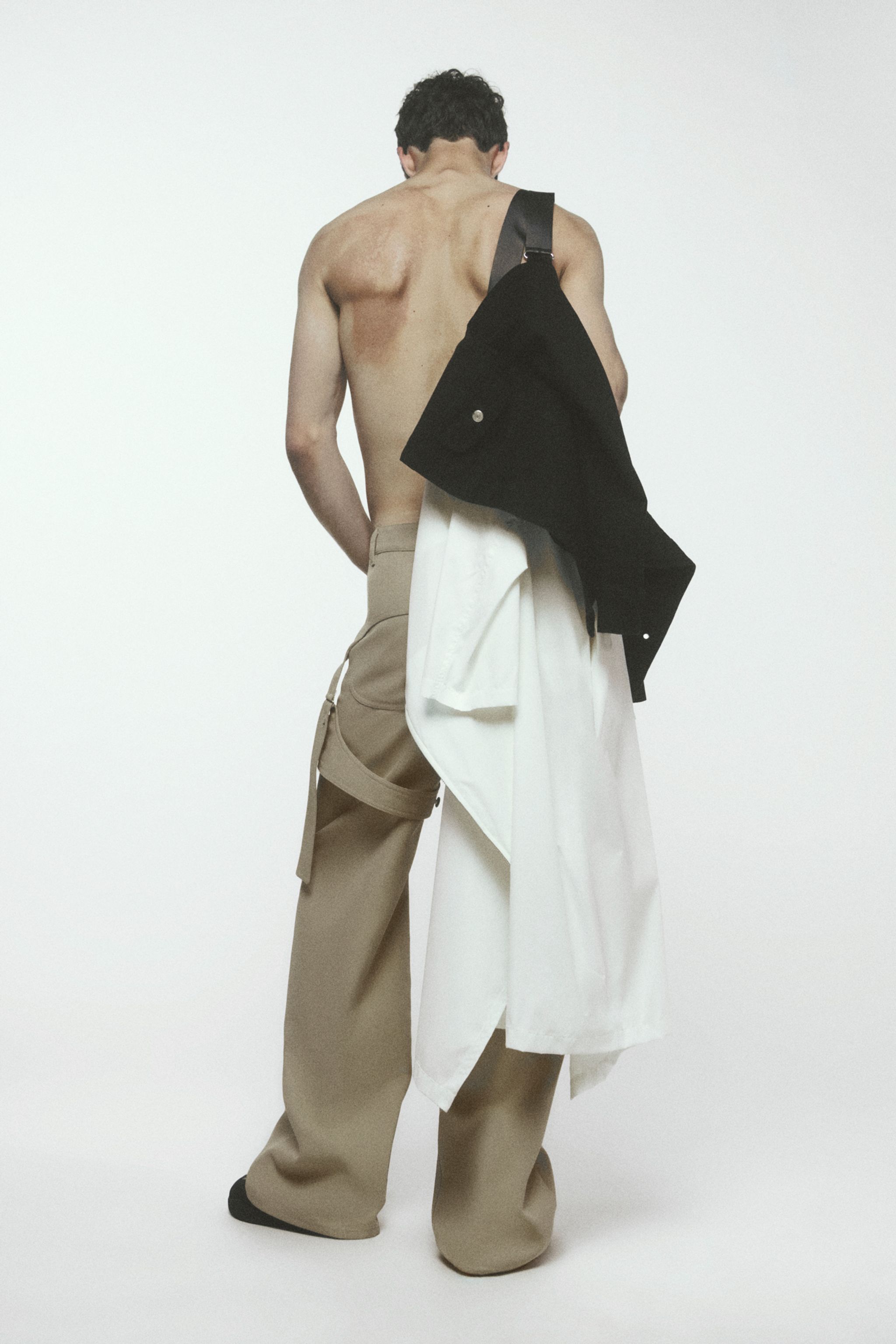 Fashion shoot featuring a shirtless male model with long beige cargo pants and a black shoulder bag.