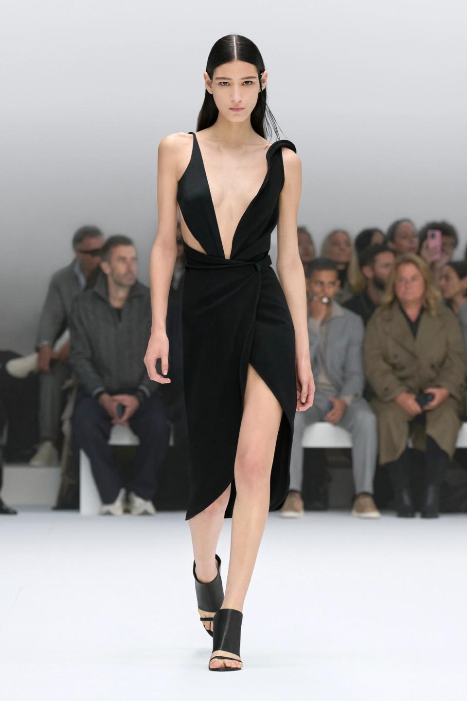 Elegant black dress with deep V neckline and high slit, showcased on a runway for Courrèges.