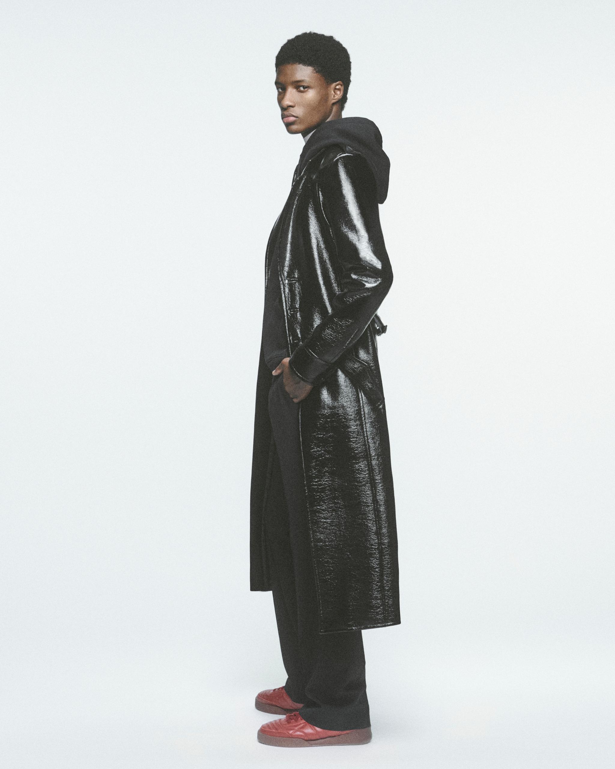 Fashion model in a glossy black long coat, hoodie, and pink sneakers for Courrèges.