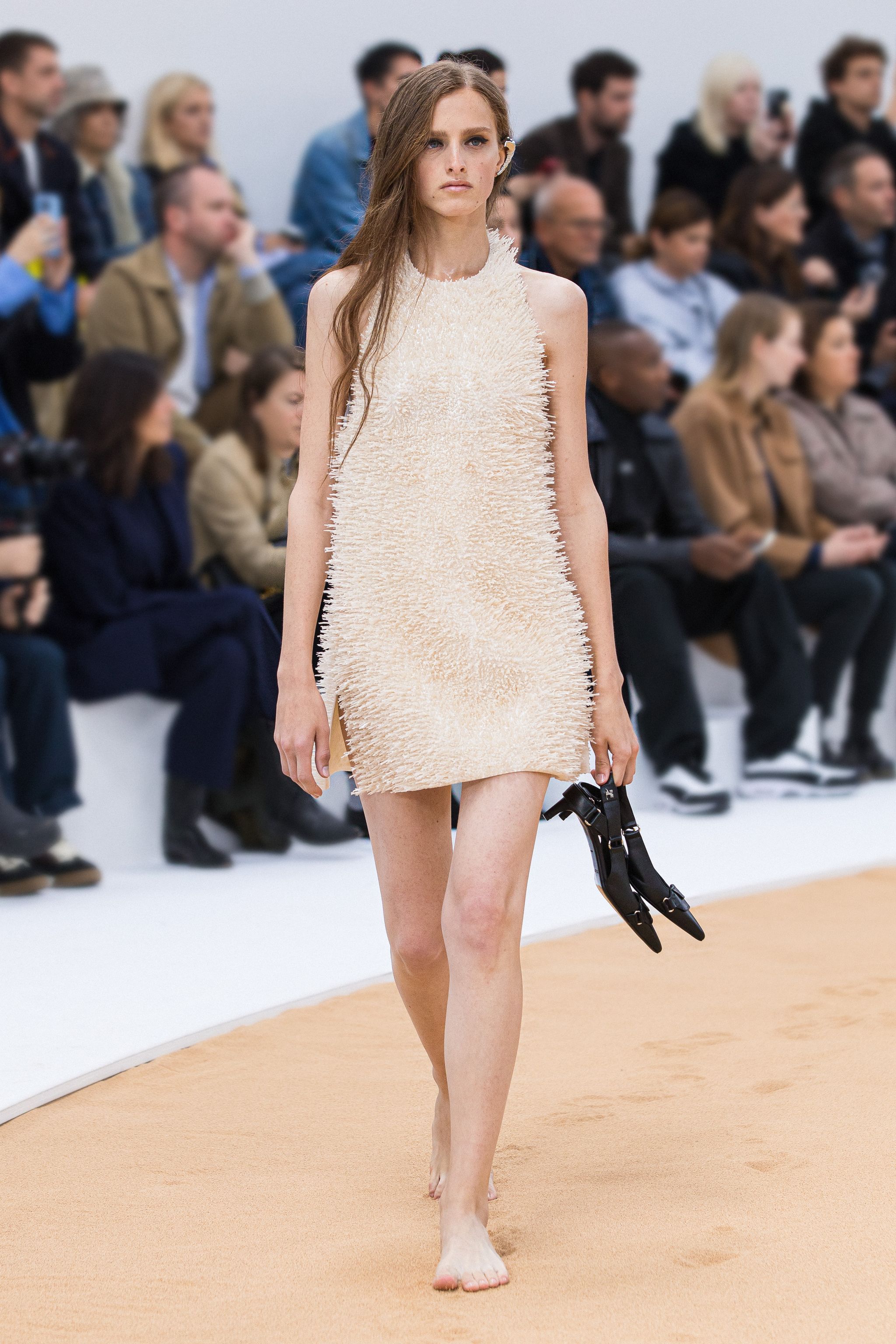 Elegant beige fuzzy dress showcased on a model during a Courrèges fashion show, with audience in background.