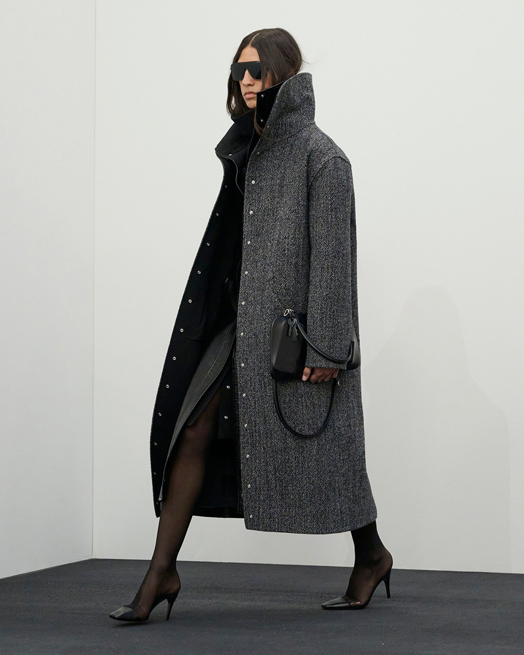 Femme élégante en long manteau gris, robe noire et talons hauts, présentant la mode Courrèges.