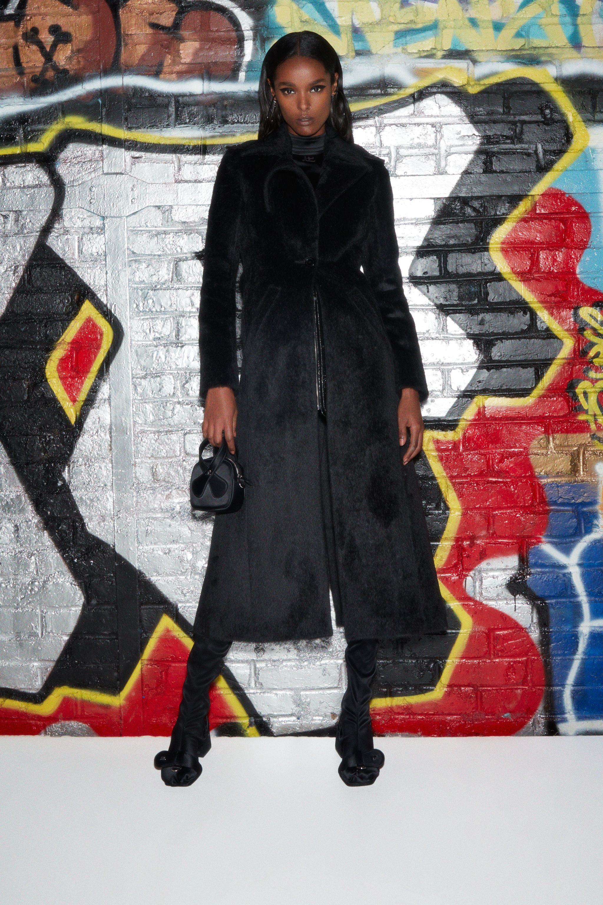 Elegant black fur coat by Courrèges displayed on a model against urban graffiti background.