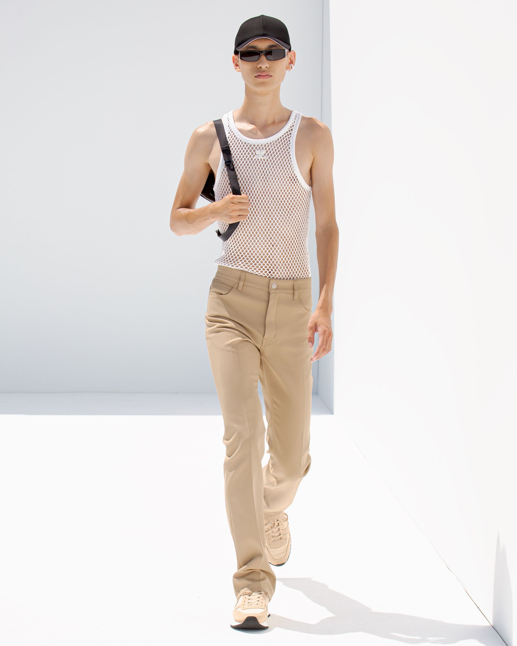 Elegant model in casual beige pants, mesh top, black cap, and sunglasses for Courrèges fashion.