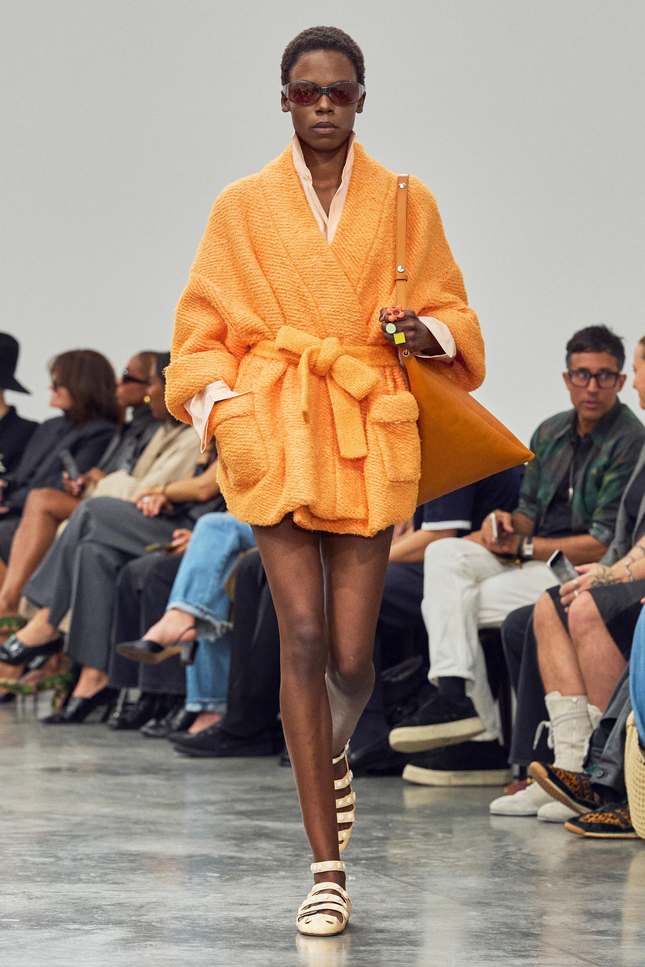 Elegant model in orange coat, sunglasses, walking on runway for Courrèges fashion show.