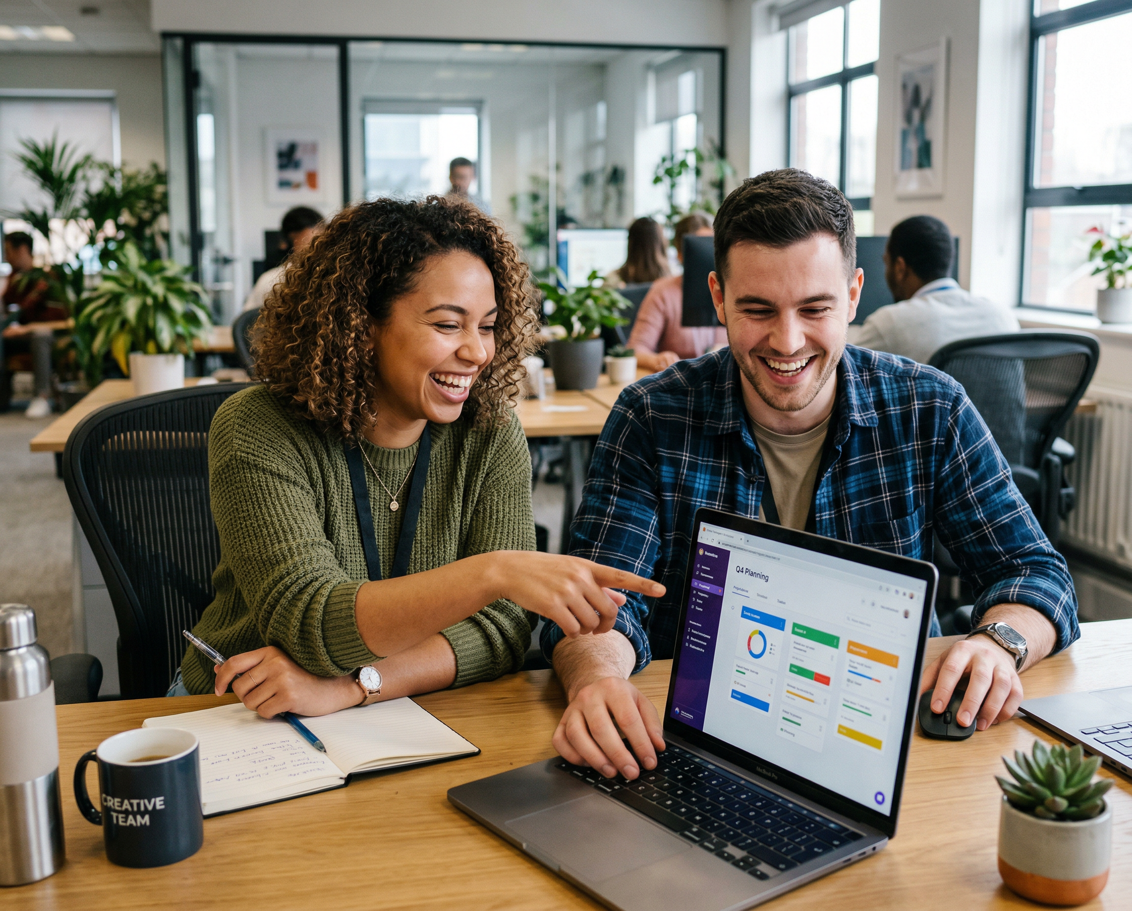 Two coworkers sitting at a desk smiling and working on a laptop