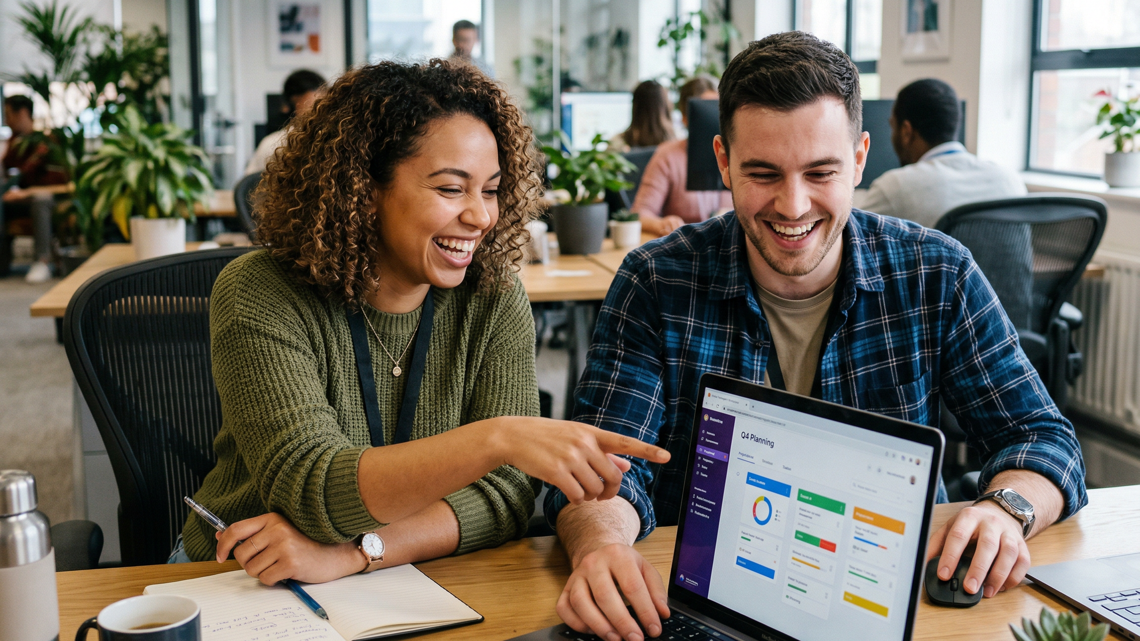 Two coworkers sitting at a desk smiling and working on a laptop