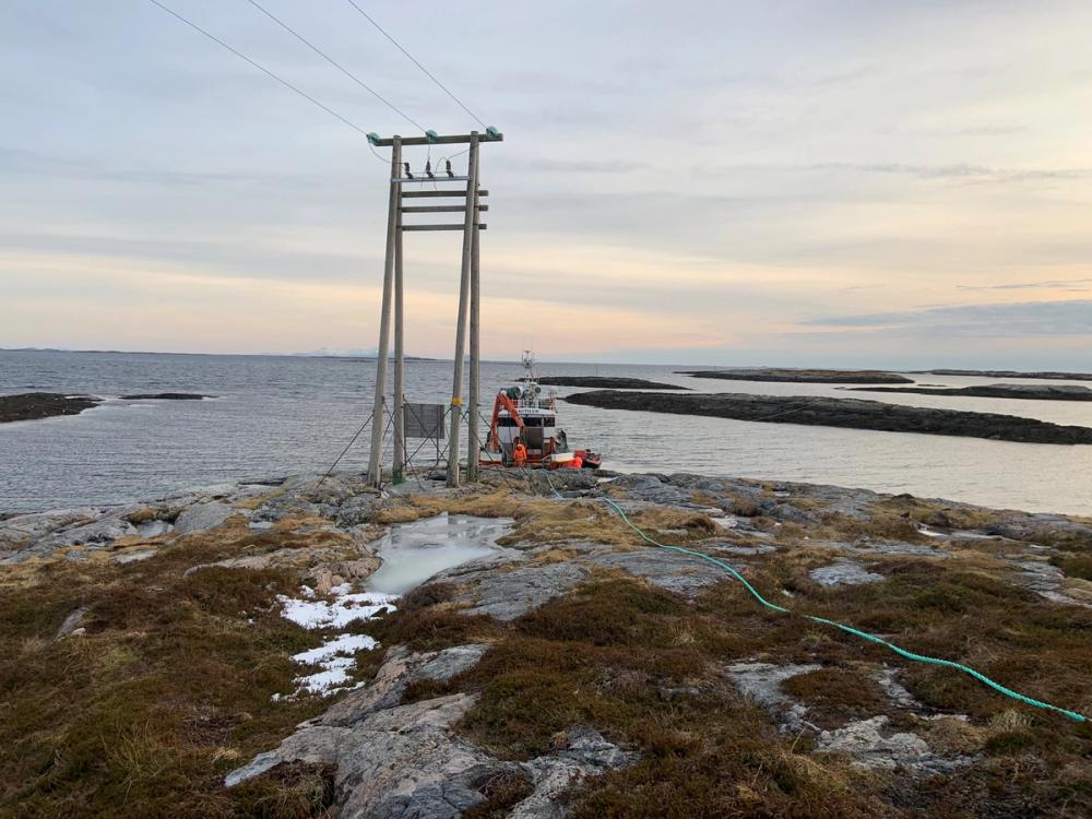 Strømstolpe på svaberget ved havet. En arbeidsbåt ligger ved land.