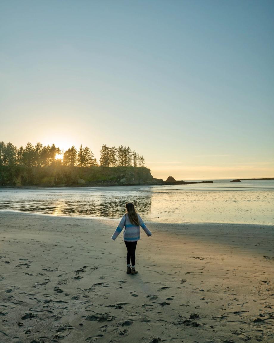 A woman is a sweater walks along a quiet beach at sundown during a visit to Sunset Bay State Park and stares at the calm water with trees in the background..