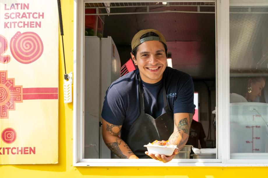 Smiling man with tattooed arms leans out the serving window of a yellow Latin Scratch Kitchen food truck, holding out a to-go container of food.