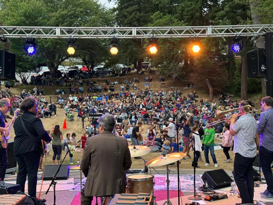 View from the stage during one of the Music on the Bay performances in Mingus Park of a live band performing with guitars, trombone, and drums, facing a large crowd seated and dancing on a grassy outdoor hillside.