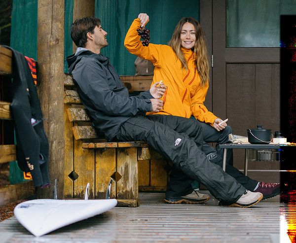 A woman and man relax on the deck of a yurt, snacking on grapes with a surfboard and camping gear nearby.