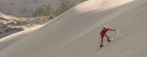 A boy in a red shirt carves down a steep sand dune on a sandboard, arm extended for balance.