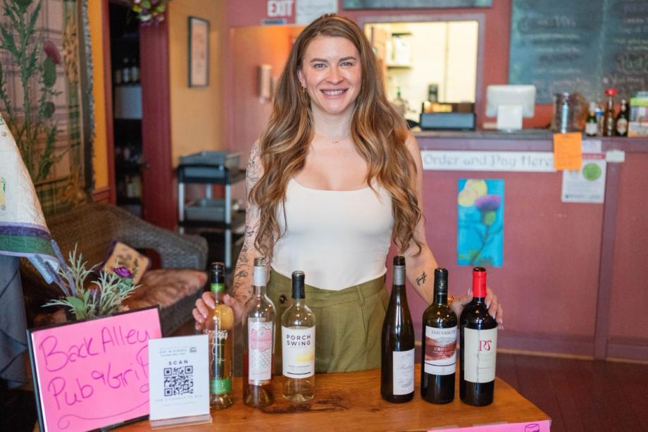 A smiling woman wearing a white tank top stands behind a table lined with wine bottles at a Back Alley Pub Grill during the North Bend Sip-N-Stroll.