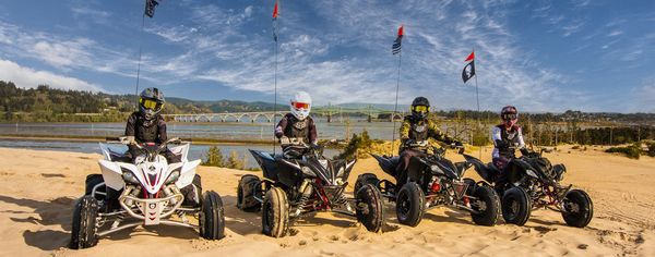 Four riders on ATVs with safety flags pause on a dune overlooking Coos Bay and the McCullough Memorial Bridge.