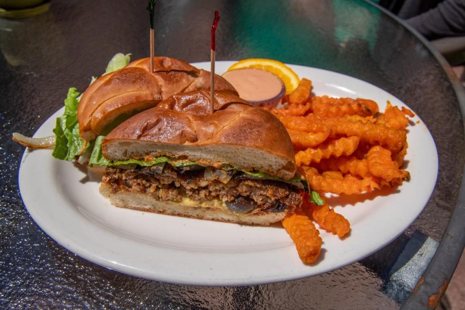 A halved vegan sandwich with lettuce and a toasted bun, served on a white plate with crinkle-cut sweet potato fries, a dipping sauce, and an orange slice.