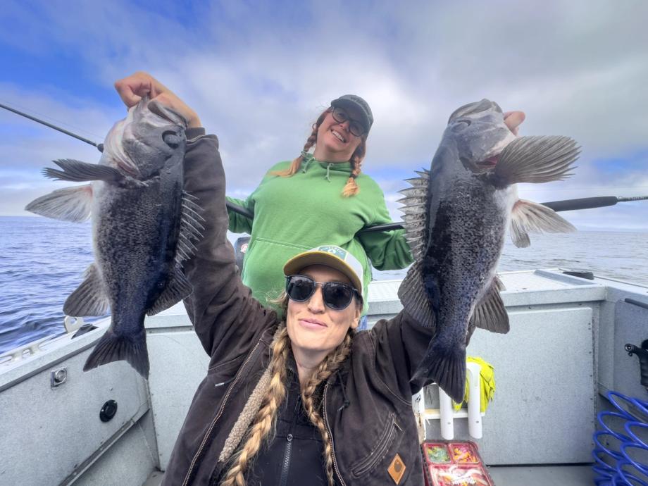 Two women on a fishing boat smiling and holding up rockfish they caught, with a cloudy sky and ocean in the background.