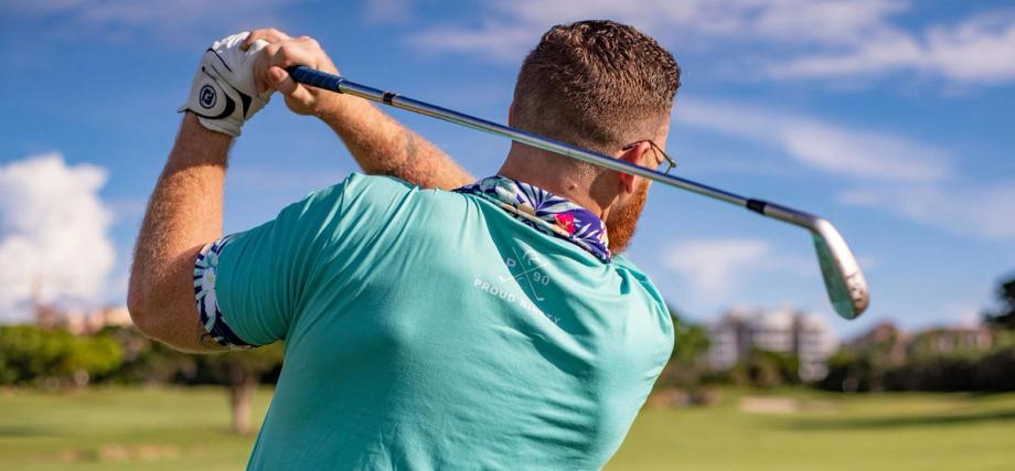 Man from behind swinging a golf club, wearing a teal Proud Ninety P90 shirt with floral trim and a golf glove.