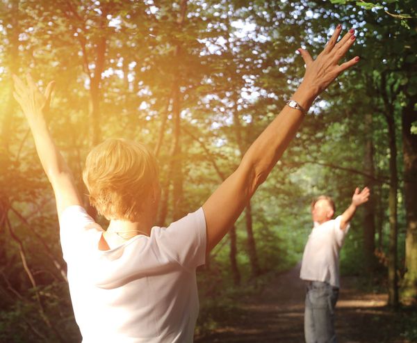 Two people standing on a sunlit forest path with arms raised, embracing the natural surroundings.