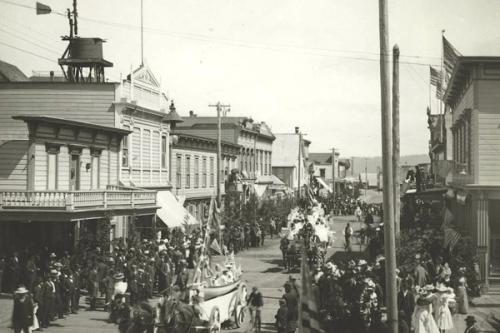 A historic black-and-white photograph of a parade along a crowded downtown Coos Bay street, with decorated floats, American flags, and Victorian-era storefronts lining the route.