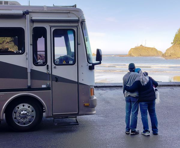 A couple stands arm in arm beside their motorhome at Sunset Bay State Park, looking out at the ocean and rocky headlands.