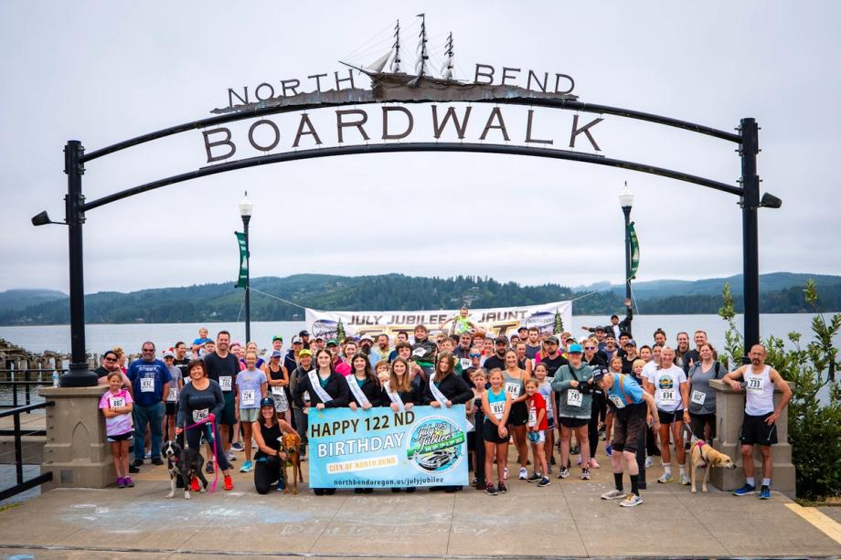 A large crowd of runners, walkers, and dogs posed for a group photo under the North Bend Boardwalk archway sign, with a bay and forested hills in the background, celebrating the city’s 122nd birthday and the 2025 July Jubilee Jaunt 5K.