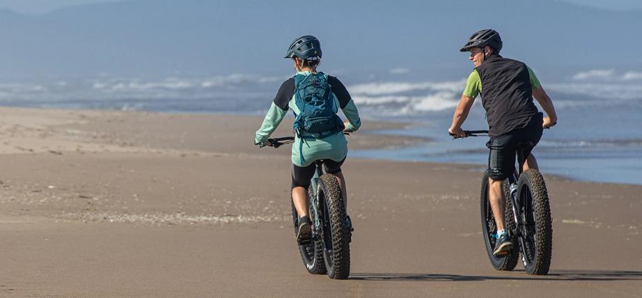 Two cyclists ride fat tire bikes along a wide sandy beach on the Oregon Coast, with ocean waves in the background.