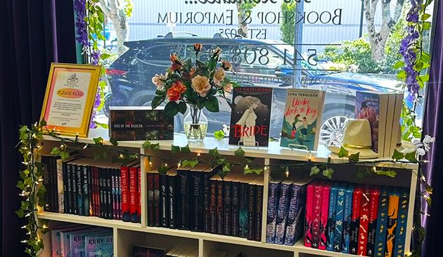 A bookshelf in a window display, featuring several books with titles like "The Bride" and "Love, Lies & Luck," a vase of flowers, a hat, and a framed sign that says "PLEASE READ"