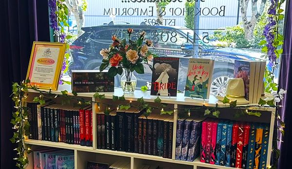 A bookshelf in a window display, featuring several books with titles like "The Bride" and "Love, Lies & Luck," a vase of flowers, a hat, and a framed sign that says "PLEASE READ"