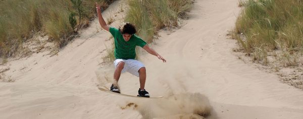 A young man on a sandboard kicks up a spray of sand after going off a jumping, arms out for balance.