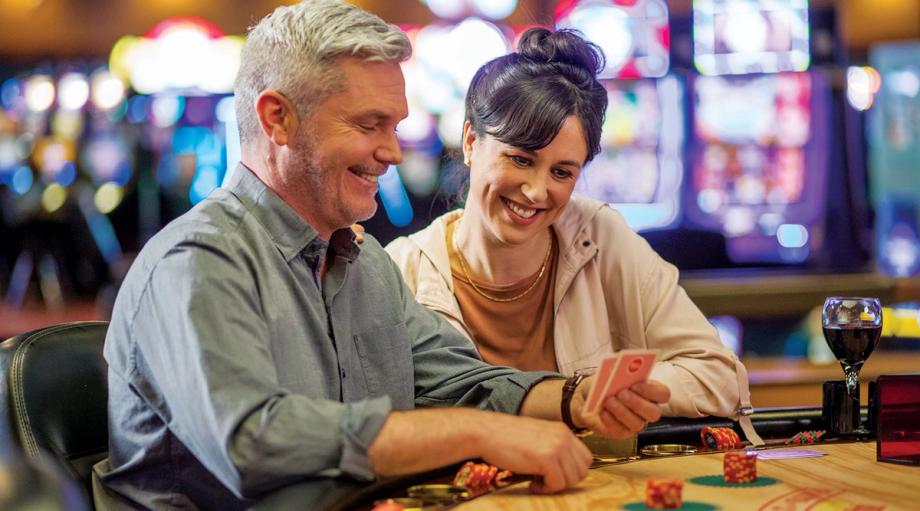 A smiling couple plays a card game at a table with poker chips and wine, with slot machines lit up in the background at Ko-Kwel Casino Resort in Coos Bay.