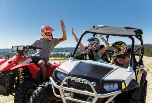 A man on an ATV high-fives passengers in an off-road buggy on a sand dune overlooking a bay, all wearing helmets.