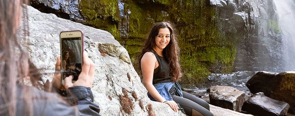 A person photographs a smiling woman seated on a large rock near the base of a mossy waterfall.