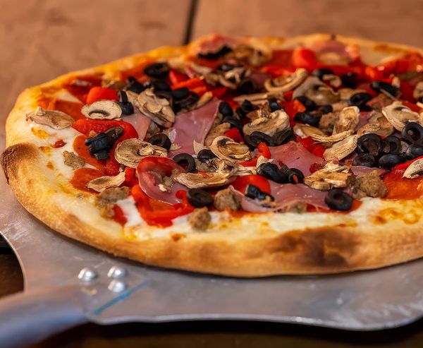 Freshly baked pizza topped with mushrooms, black olives, red bell peppers, and prosciutto on a metal pizza peel.