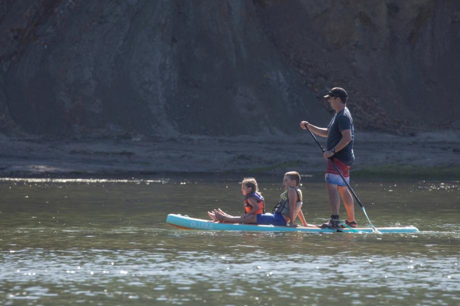 Man stands paddling a stand-up paddleboard in the calm waters below a steep rocky bluff at Sunset Bay State Park in Coos Bay while two children in life vests sit in front of him.