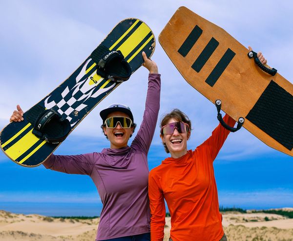 Two smiling women in sun gear hold sandboards overhead, standing on a dune above the ocean.