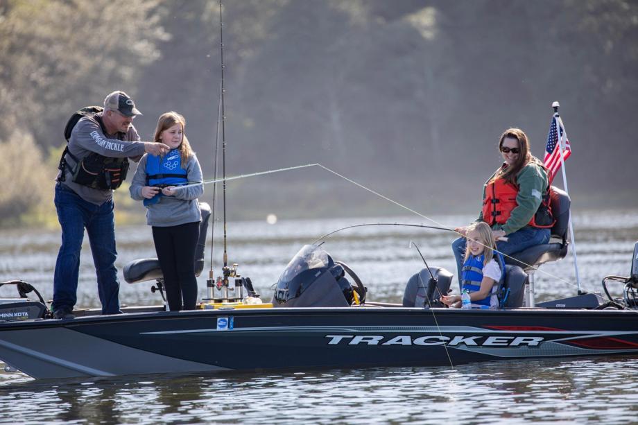 A family fishing together on a boat, with an adult helping a girl hold her fishing rod as another woman and child fish nearby, an American flag flying at the stern.