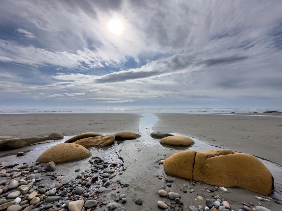 A wide, flat beach at low tide on the Oregon Coast, with smooth rounded boulders and scattered pebbles in the foreground. A narrow stream of water flows between the rocks toward the ocean in the distance. The sky is overcast with dramatic clouds, and the sun breaks through near the center, casting a soft glow over the scene.