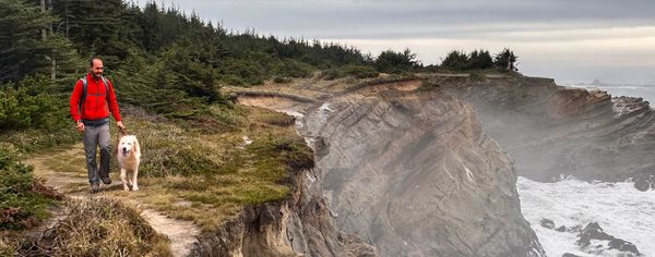 A man in a red jacket walks his dog on a leash along a coastal bluff trail on the Sunset Trail, with cliffs and ocean below.