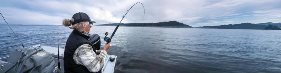 alt="A woman reels in a fish from the stern of a charter boat on the Pacific Ocean off the Oregon Coast.