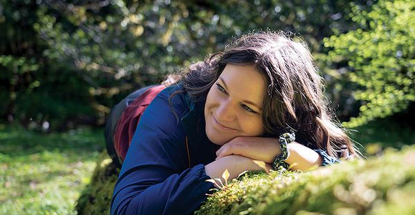 A woman smiling and resting her chin on her hands on a moss-covered log in a lush green forest