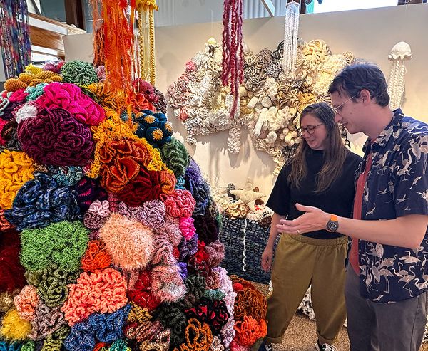 Two visitors admire a large-scale crocheted coral reef installation at the Coos Art Museum, with vivid colors and textile sea creatures filling the gallery space.