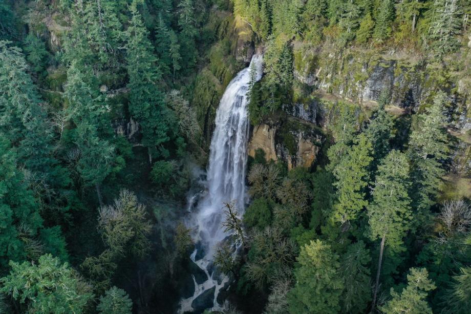 An aerial view of Silver Falls in Coos County, Oregon, with a tall waterfall cascading down a mossy rock face into a misty pool, surrounded by dense evergreen forest.