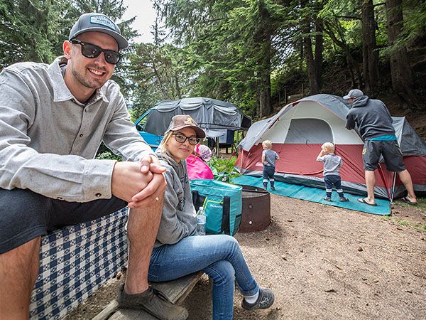 A family sets up camp among towering trees at Sunset Bay Campground on the Oregon Coast, with two adults relaxing near a fire pit and children helping pitch a tent.