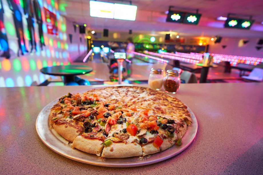 A large sliced pizza topped with tomatoes, olives, green peppers, and mushrooms, on a plate at a bowling alley counter with salt and pepper shakers, colorful disco lighting, and bowling lanes visible in the background.