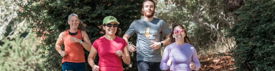 Four smiling runners jog together on a wooded trail, one wearing a Wild Coast Running Co. shirt.