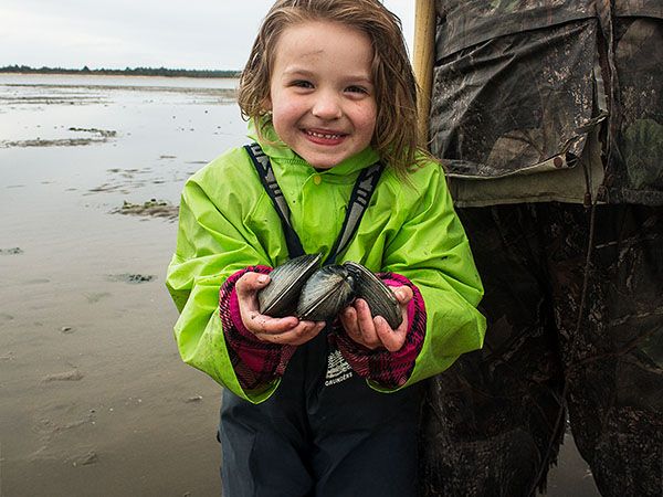 A young girl in a bright green rain jacket smiles while holding freshly dug clams on a muddy tidal flat