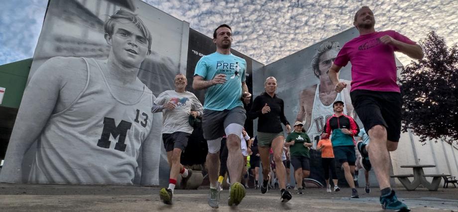 A group of runners jogs past the Steve Prefontaine mural in downtown Coos Bay, one wearing a Prefontaine 10K Run shirt.