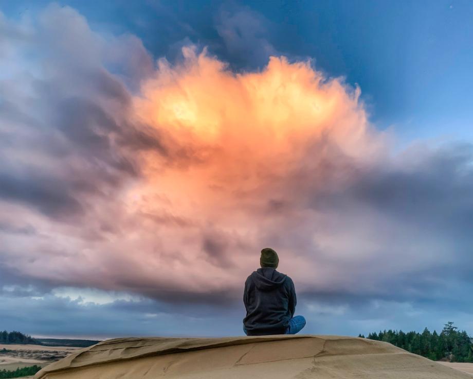 A person sits alone on top of a sand dune at the Oregon Dunes, gazing up at a dramatic sky with a large, glowing golden cloud formation surrounded by sweeping storm clouds at sunset.