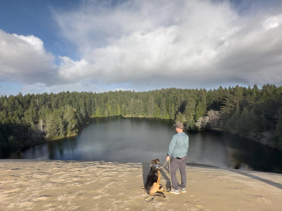 A man in a teal jacket and a leashed black and tan dog sitting together at the edge of a sandy dune, looking out over a calm, tree-ringed lake under a partly cloudy sky.